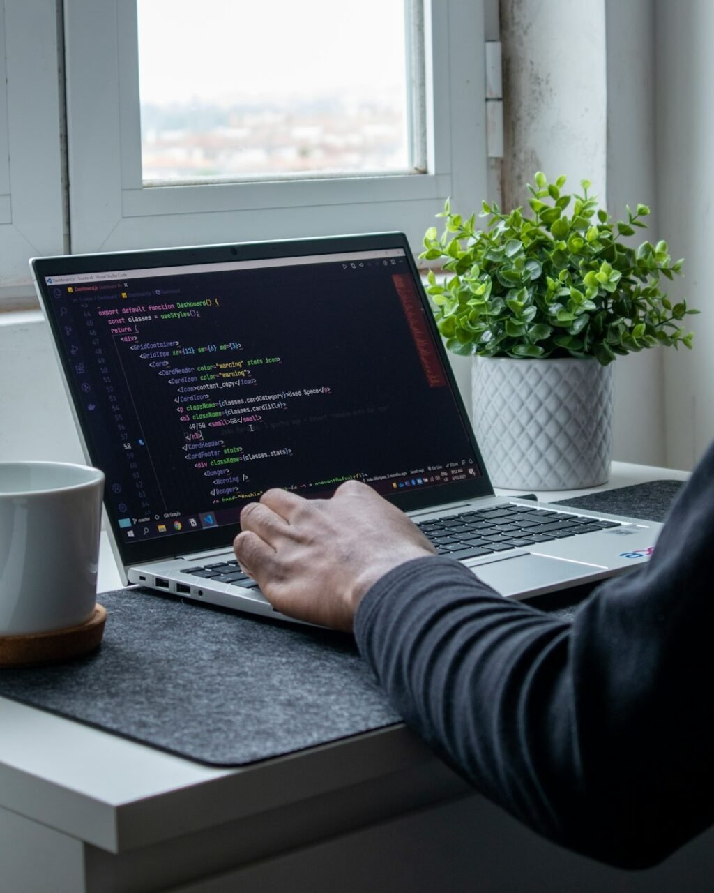 Web developer typing code on a laptop beside a plant and mug